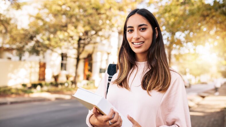 Young hispanic female student with book in college campus. Beautiful young woman with book on the road.