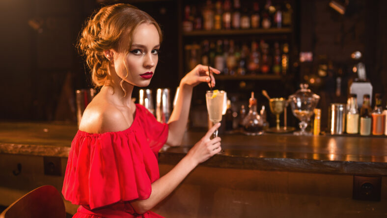 Beautiful woman in red dress drinks alcohol cocktail at the bar counter. Date in nightclub, attractive love couple in pub