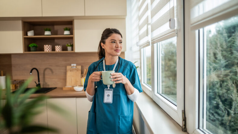 Female medical professional in blue scrubs holding a cup of coffee, pausing to look out a window in a modern kitchen, reflecting on her work during a well-deserved break