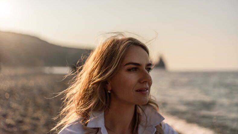Close-up of a beautiful blonde woman with wind-swept hair, looking away thoughtfully at the serene ocean during a golden sunset on a pebble beach.
