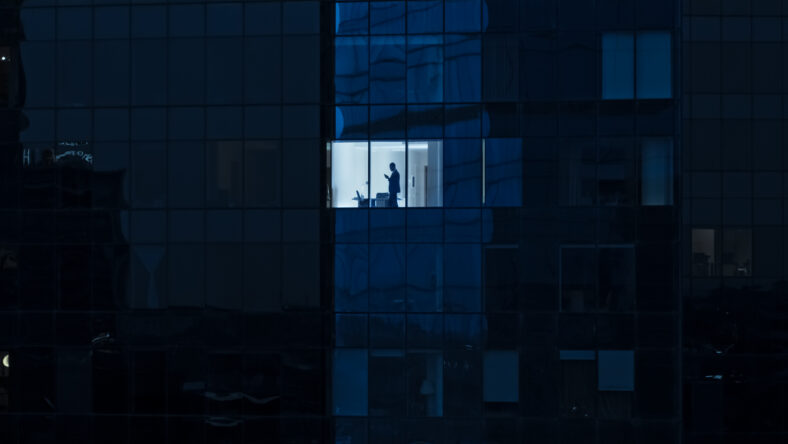 Aerial Shot: From Outside into Office Building with Businessman Using Mobile Phone and Standing by the Office Window. Beautiful Shot of The Financial Business District Skyscrapers.