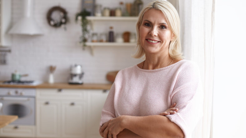Attractive middle aged housewife with blonde hair and brown eyes posing indoors in her modern clean stylish kitchen interior, crossing arms on her chest and looking at camera with pleased smile