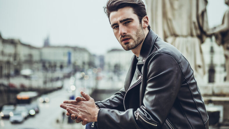 One handsome young man in urban setting in modern city, standing, wearing black leather jacket and jeans, looking at camera