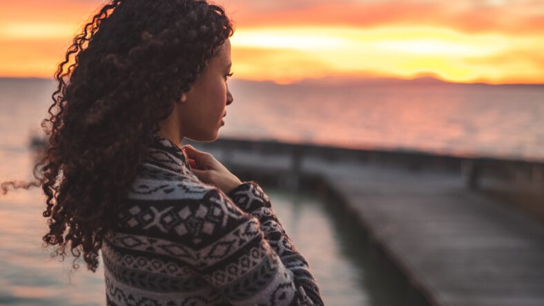Thoughtful evening mood with a young Afro American woman standing on the promenade by the lake and looking towards the water and the setting sun. Burning sky
