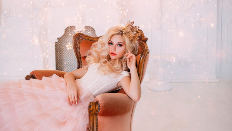 Young beautiful queen woman resting in medieval armchair. beauty face. New Year garland scenery glare sparks bright. Backdrop white classic room interior. Hairstyle princess girl blonde long wavy hair
