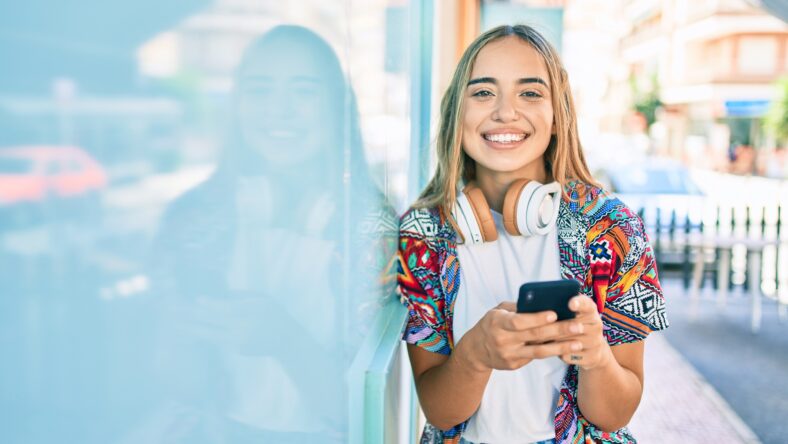 Young beautiful blonde caucasian woman smiling happy outdoors on a sunny day wearing headphones and using smartphone