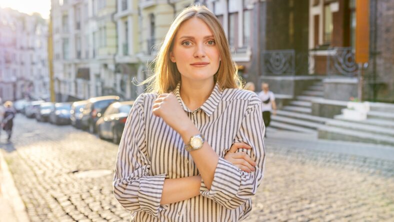 Urban portrait of beautiful young blonde woman on city street at sunset. Smiling happy female looking at camera