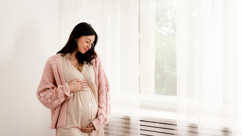 Young beautiful woman on second trimester of pregnancy. Close up of pregnant female in pink oversized knitted sweater with arms on her round belly. Expecting a child concept. Background, copy space.