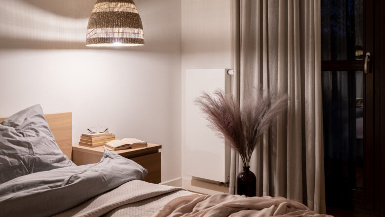 Stylish bedroom with wooden bedside table, wicker lamp and decorative feathers in vase next to comfortable bed
