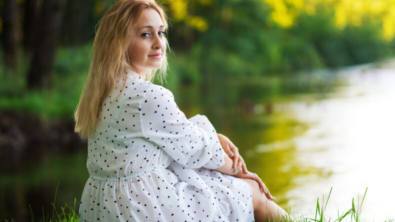 Beautiful young blonde woman in a white dress on the shore of a lake or river on a sunny day. Enjoy your life