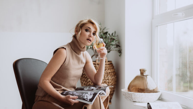 Young businesswoman works at home. Pensive calm model holds a glass of white wine and a magazine and looks at the camera. Beautiful woman in a modern interior. Soft selective focus.