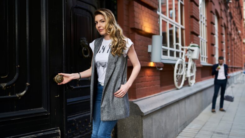 Half length portrait of Caucasian female tourist opening vintage doors in historic center during travel vacations for visiting ancient city, young woman in casual clothes looking at camera