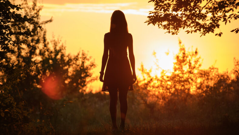 Back view of young woman in summer dress walking alone through evening dark forest.