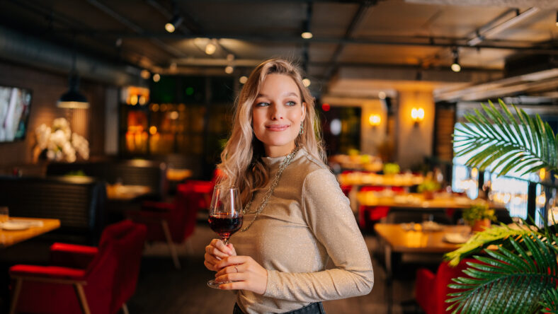 Portrait of attractive female holding glass of red wine standing in restaurant with luxury dark interior, looking away. Front view of cute woman posing at cafe, blurred background.