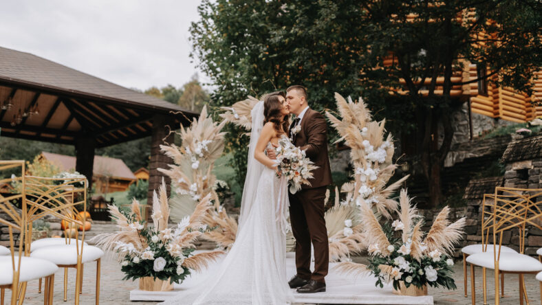 Groom and bride kiss. Newlyweds with a wedding bouquet standing at a wedding ceremony under an arch decorated with flowers and dried flowers outdoors.