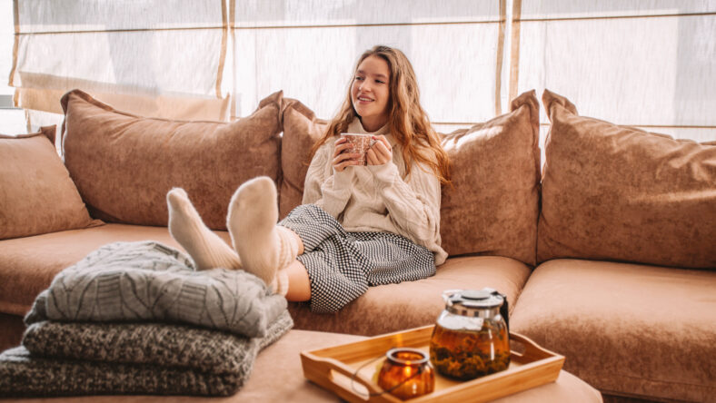 Young smiling woman sitting on sofa and looking up while drinking hot tea. Young millennial woman thinking at home in leisure time. Happy girl relaxing at home on bright winter morning