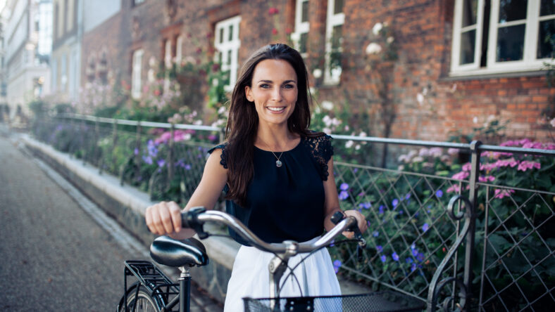 Beautiful woman with a cycle walking down the city road. Caucasian female smiling and looking at camera.