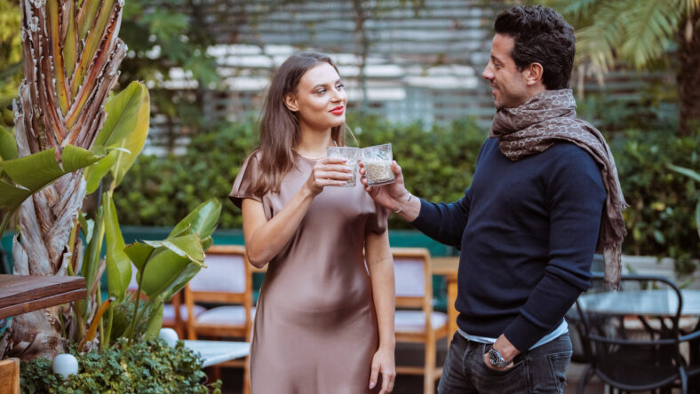 Smiling beautiful young couple toasting and chatting at terrace of a modern bar with tropical plants, drinking cocktails. Human relationships, dating concept.