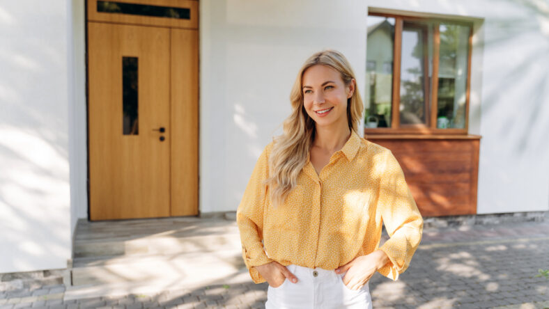 Blonde real estate agent smiling confidently while posing in front of a modern new house, embodying success in the property market