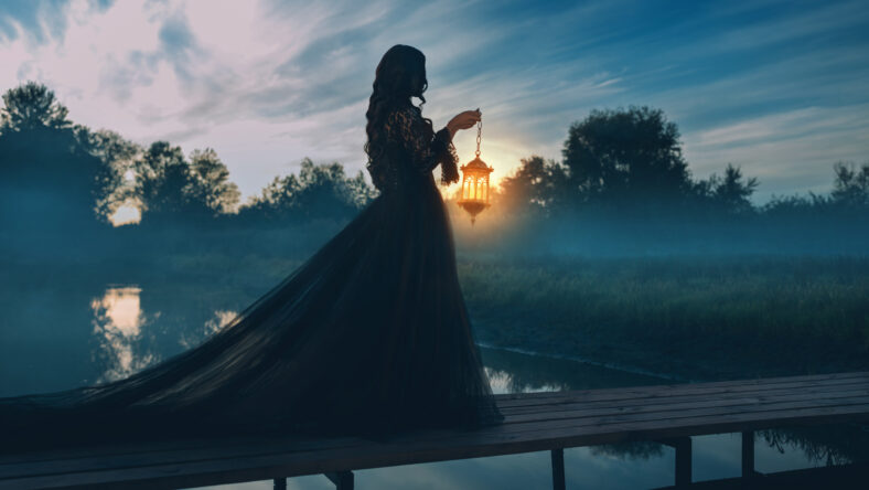 photo real person silhouette gothic woman hand hold lantern lamp walking across bridge over river dense fog mist night nature dark dramatic blue sky lake. long black dress back rear view fantasy girl