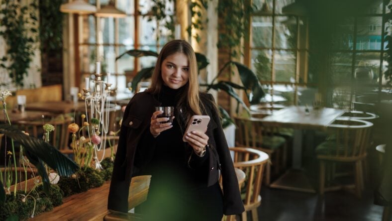 Beautiful young woman standing with smartphone and glass of drink in cafe