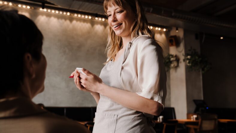 Positive waitress writes down order of mature woman at table in cafe