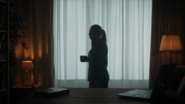 Silhouette of a woman with a phone to her ear and a cup of coffee in her hand. There is a diary on the table next to the laptop