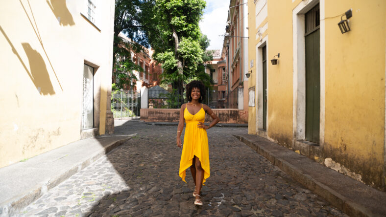 Beautiful young black woman walking on the cobblestone streets wearing yellow clothes, with afro hair, smiling and happy. Confident person on a trip
