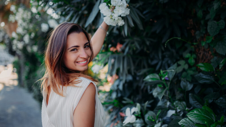 A beautiful brunette girl stands in a white dress on the background of bushes and flowers