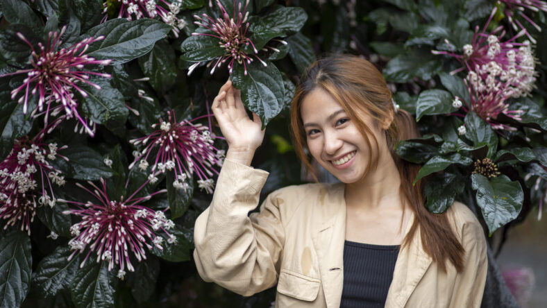 Beautiful smiling Asian woman feeling happy in an outdoor garden. She stands near natural plant with purple flowers, enjoying serene moment in nature