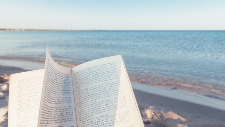 Relax concept. Open book on sand near the sea. Summer vacations still life. The text is transformed and not recognizable.