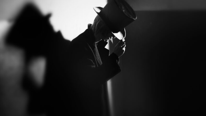 Shadowgraph of the man in black coat and hat standing near the wall and his shadow. Monochrome photo with natural darkness. Artistic grain added for movie effect