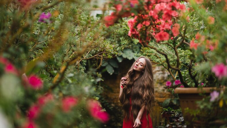 Young beautiful caucasian woman in glass greenhouse among colorful azalea flowers. Art portrait of a long-haired girl wearing a romantic red dress.