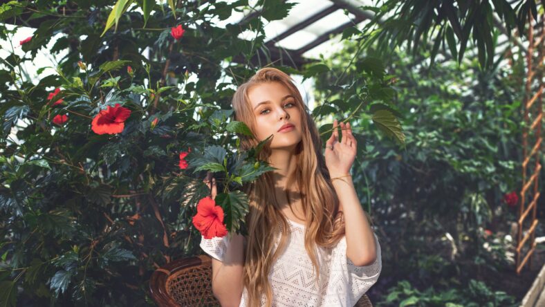 Young beautiful happy blonde girl model with red flowers on tropical plants background in greenhouse