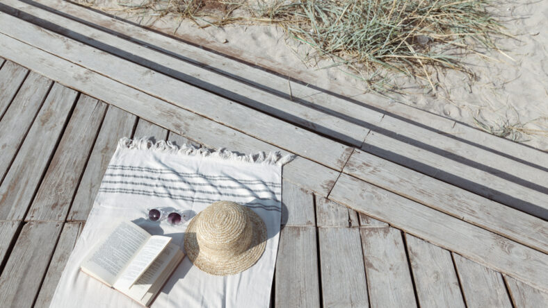 Boho eco styled beach essentials on sand by sea. Straw hat, glasses, book and beach towel on wooden backround, top view.