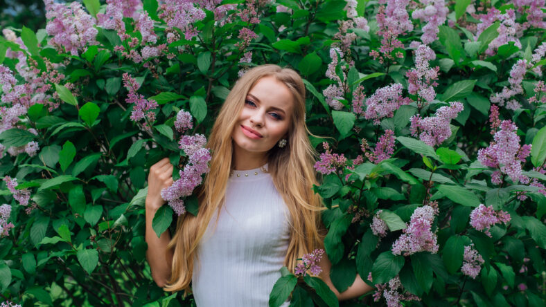 Spring portrait of a charming blond woman wearing beautiful white dress standing next to blooming purple lilac bush.