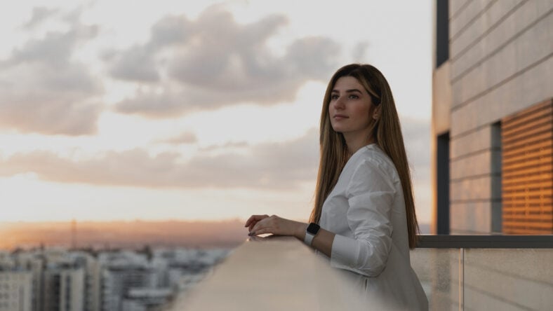 Young pretty woman dressed on white shirt looking at city from balcony, enjoying the sunset at terrace. Shot of beautiful young woman standing on the balcony and looking at the view. Relax, dreaming