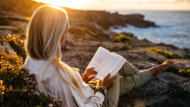 A Hispanic female sitting at the rocky beach reading a book at sunset