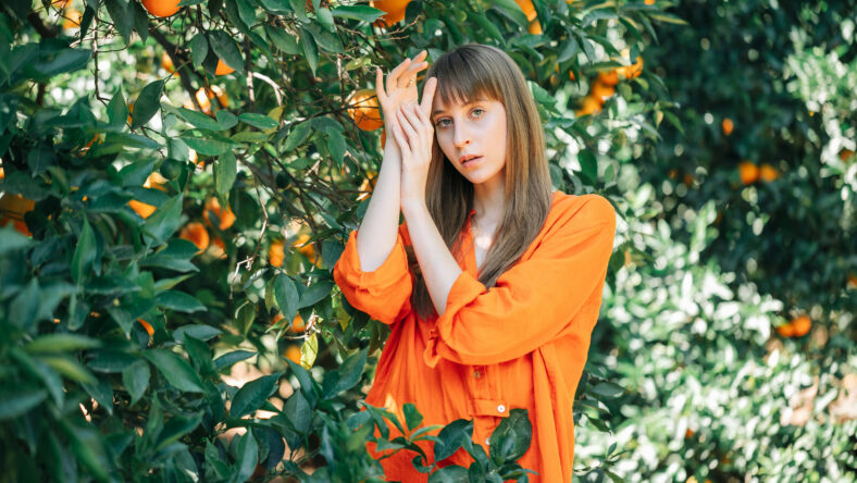Seriously girl in orange dress is posing to camera by holding hands up together in orange garden