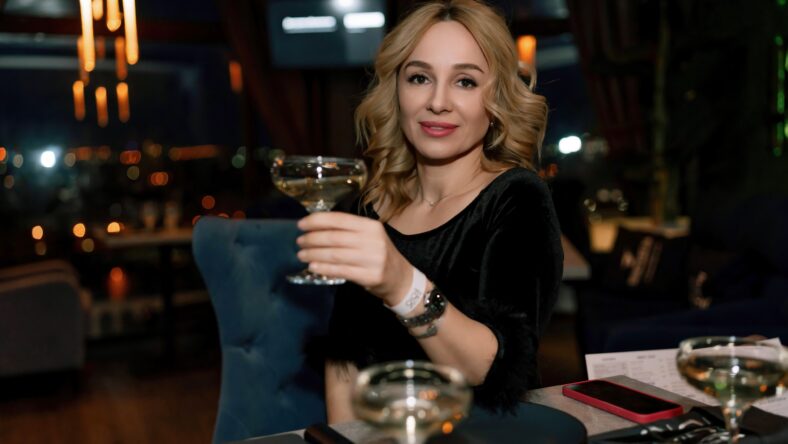 woman in a restaurant with champagne in her hand. She is sitting at a table against the backdrop of a dark restaurant hall with lamps