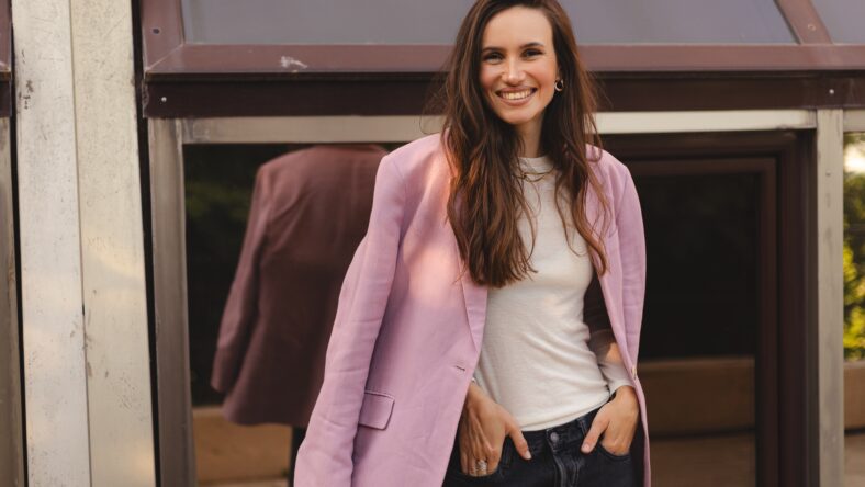 Stylish young woman smiling outdoors, wearing a pink blazer over a casual white t-shirt and black jeans. Confident modern look, street style fashion, candid urban portrait.