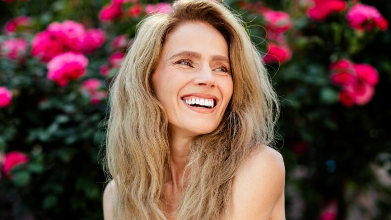 A charming middle-aged woman with radiant blonde hair smiling against a backdrop of vivid pink roses under glowing natural sunlight, exuding joy and serenity
