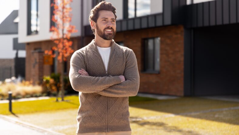 Young man smiling with arms crossed, standing confidently in front of his new suburban home