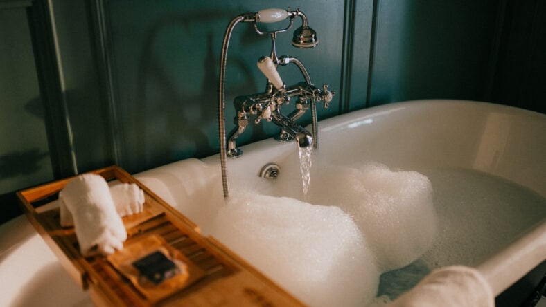 Luxurious clawfoot bathtub filled with bubbly water, set against dark teal walls. Wooden tray holds rolled towels and a soap dish beside the tub. The vintage-style chrome faucet drips gently