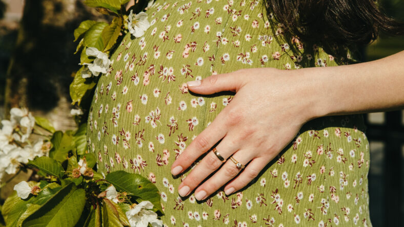 An unrecognizable Caucasian girl in a green dress holds her stomach with one hand while standing between the branches of a blossoming apple tree in the front yard of a house on a sunny spring day in the countryside. A side view, very close-up. Concept: expecting a child, happy motherhood.