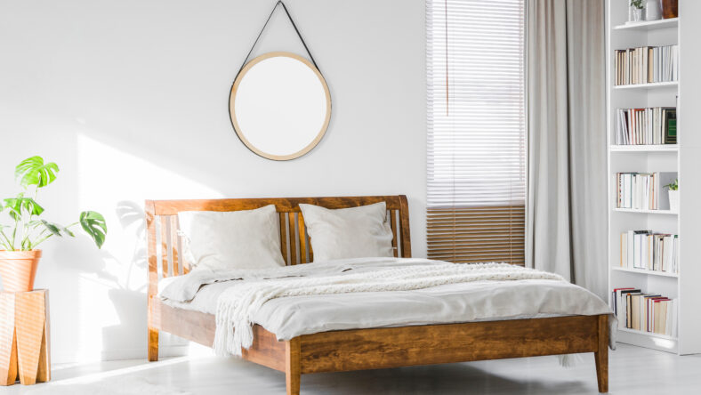 A wooden rustic bed frame and a home library bookcase in a natural, sunlit hotel room interior with white walls