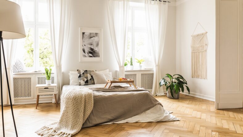 Framed poster on a white wall above a cozy double bed with beige sheets and breakfast tray in a spacious, sunny bedroom interior