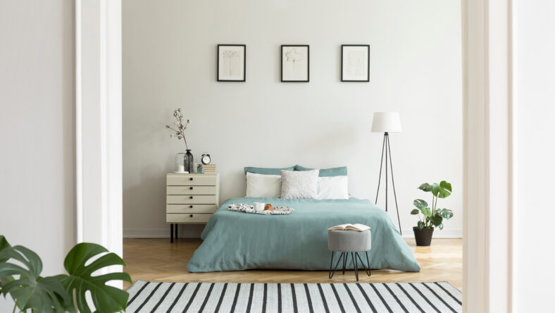 A view from a different room into a pastel bedroom interior with a big bed in the middle and a lamp and a night cabinet beside. Real photo.