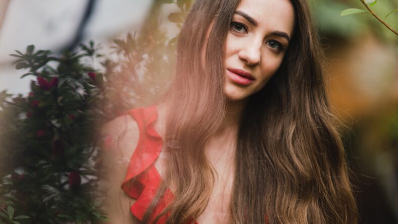 Young beautiful caucasian woman in glass greenhouse among colorful azalea flowers. Art portrait of a long-haired girl wearing a romantic red dress.