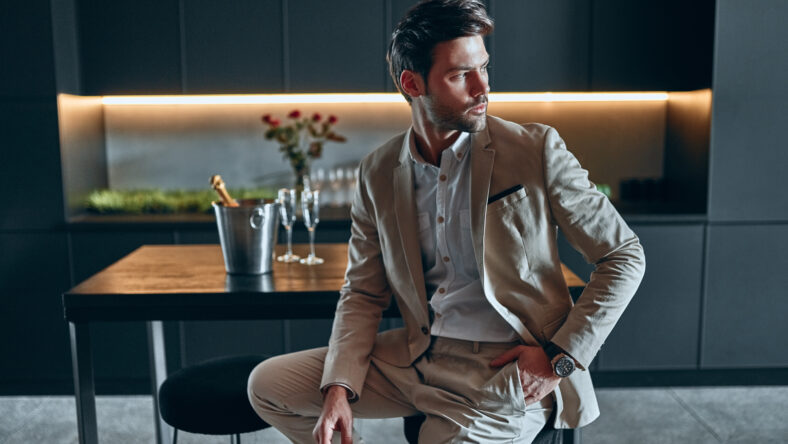Handsome stylish man in beige suit at home. Sitting in modern black kitchen.